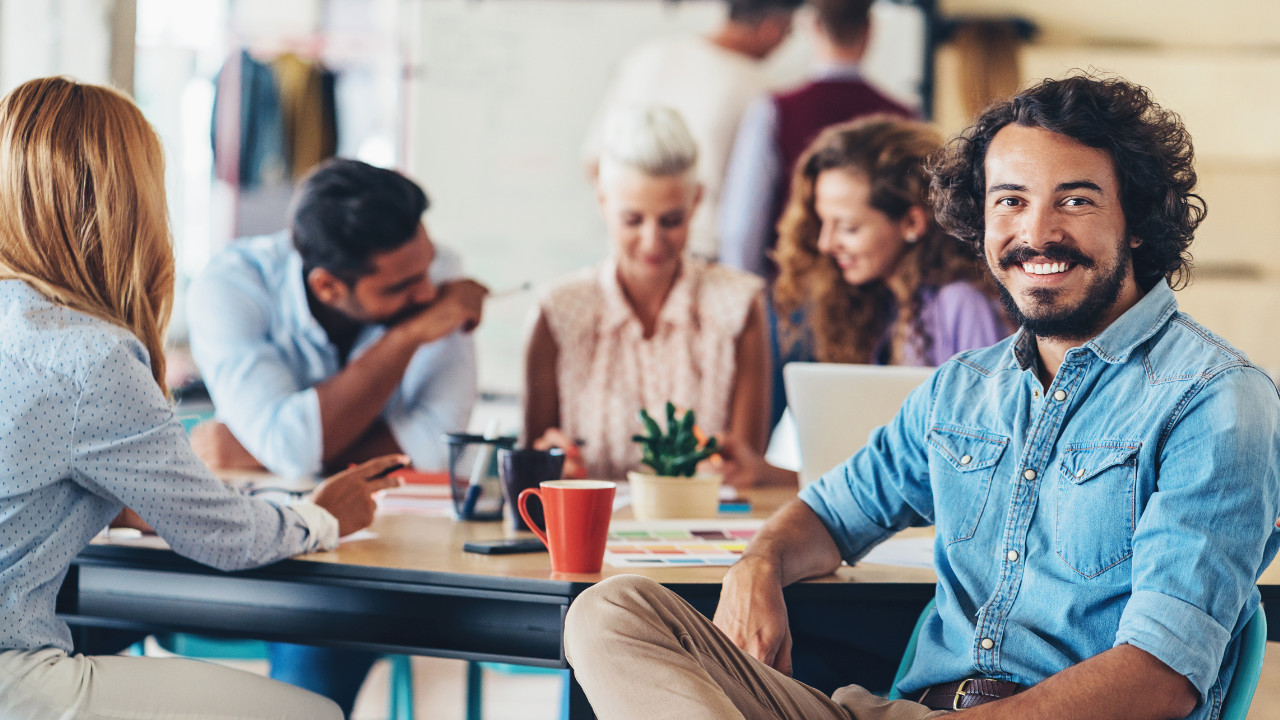 A smiling man sits at the end of a boardroom table of professionals in a meeting